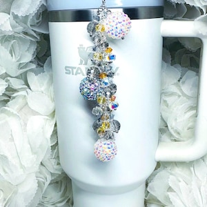 May include: A white tumbler with a beaded charm. The charm features multiple crystal beads in clear, yellow, and iridescent colors, with a large, sparkly ball at each end. The tumbler has a white handle and a light blue rim.