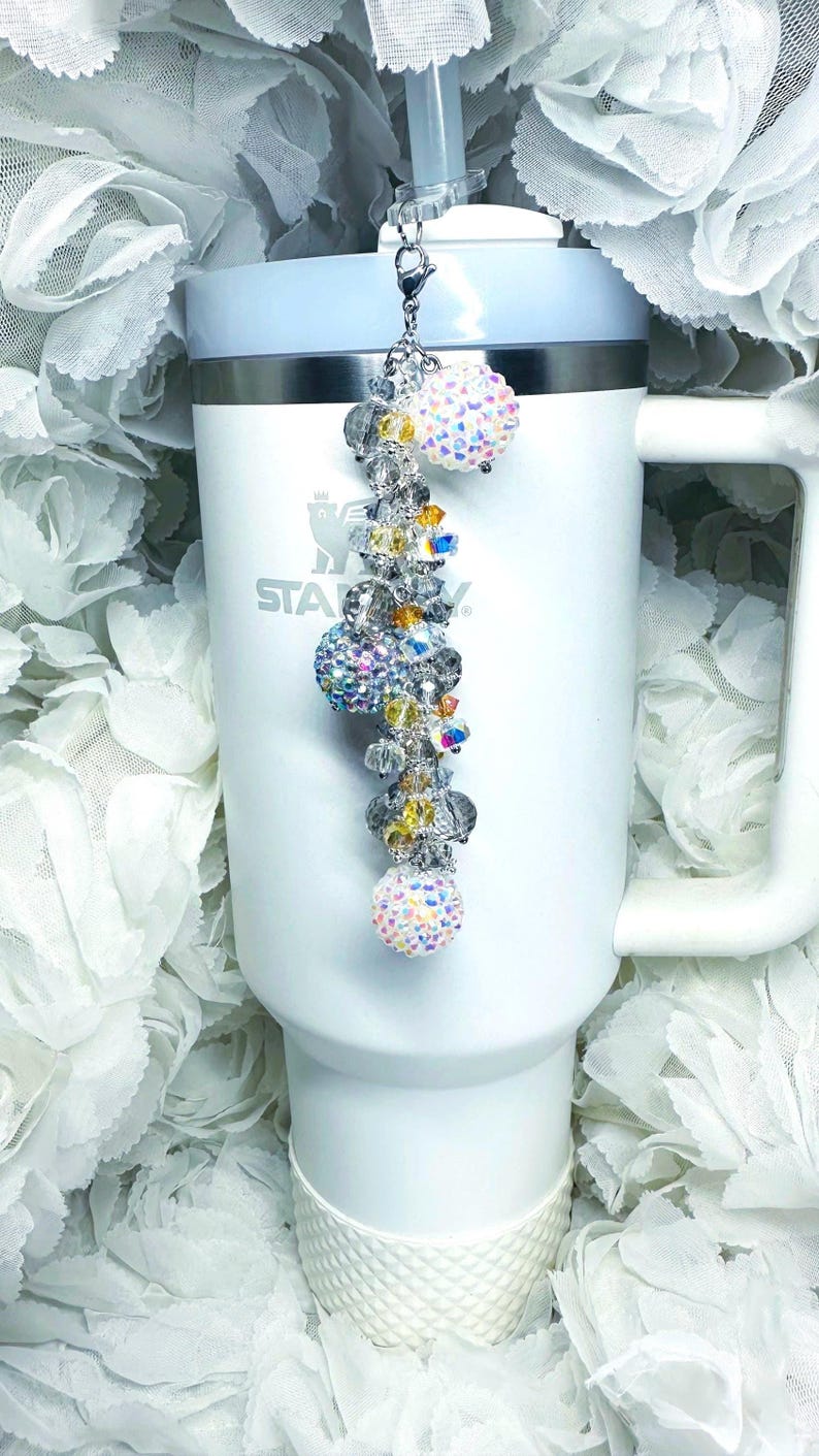 May include: A white tumbler with a beaded charm. The charm features multiple crystal beads in clear, yellow, and iridescent colors, with a large, sparkly ball at each end. The tumbler has a white handle and a light blue rim.