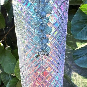 May include: A tall, iridescent tumbler with a diamond-cut pattern. The cup is adorned with a beaded charm in shades of blue and silver. The background features green leaves, creating a contrast with the cup's shimmering surface.