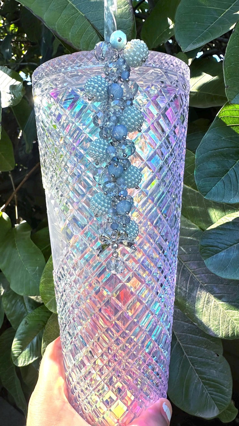 May include: A tall, iridescent tumbler with a diamond-cut pattern. The cup is adorned with a beaded charm in shades of blue and silver. The background features green leaves, creating a contrast with the cup's shimmering surface.