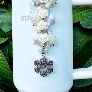 May include: A white tumbler with a decorative charm. The charm features white pearl beads, iridescent orbs, and white rose accents, topped with a silver-colored filigree flower. The tumbler has the word "STA" printed on it.