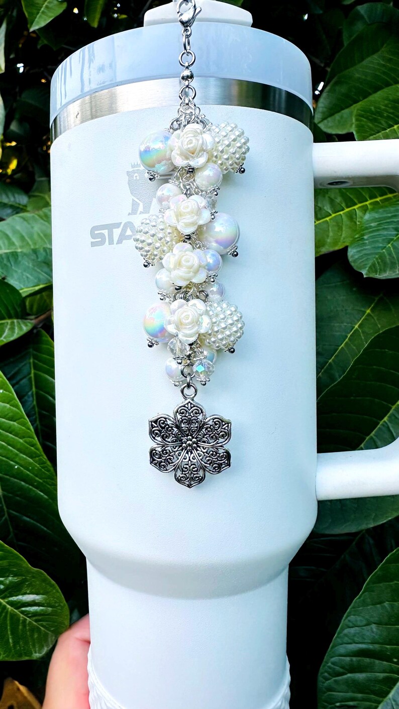 May include: A white tumbler with a decorative charm. The charm features white pearl beads, iridescent orbs, and white rose accents, topped with a silver-colored filigree flower. The tumbler has the word "STA" printed on it.