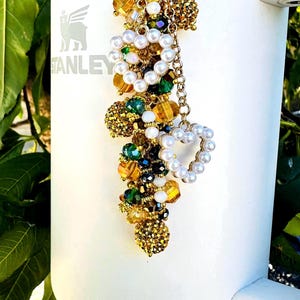 May include: A white Stanley tumbler with a decorative charm. The charm features a gold chain with clusters of beads in gold, green, and clear colors, along with a heart-shaped pearl accent. The tumbler has the Stanley logo.