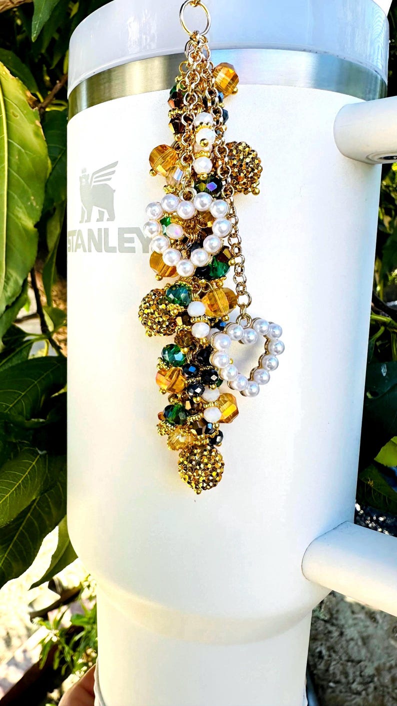 May include: A white Stanley tumbler with a decorative charm. The charm features a gold chain with clusters of beads in gold, green, and clear colors, along with a heart-shaped pearl accent. The tumbler has the Stanley logo.