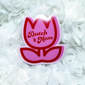 May include: A pink tulip-shaped sticker with the words "Dutch Mom" in red script. The sticker has a white border and a small windmill graphic. The background is a white, textured fabric.