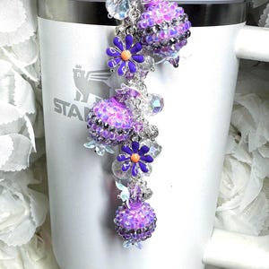 May include: A decorative charm for a tumbler, featuring a cascade of purple and silver beads, crystals, and floral accents. The charm is attached to a white tumbler with the word "STANLEY" visible.