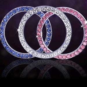 May include: Three interlocking silver rings, each encrusted with sparkling crystals. One ring features blue crystals, another white, and the third pink. The rings overlap, creating a visually appealing design against a dark background.