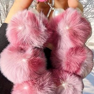 May include: Pink fluffy keychain accessories. The keychains are made of soft, plush material in varying shades of pink. Each keychain has a silver-colored chain and a pink ring for attaching keys. A white pearl-like bead hangs from the chain.