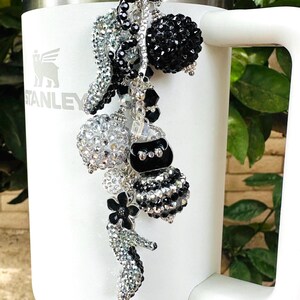 May include: A white Stanley tumbler with a decorative charm. The charm features various black and silver beaded elements, including a purse, flower, and spheres. The charm is attached to the tumbler with a silver heart-shaped clasp.