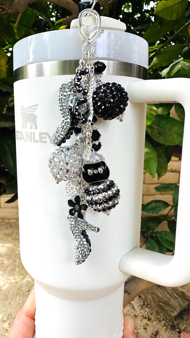 May include: A white Stanley tumbler with a decorative charm. The charm features various black and silver beaded elements, including a purse, flower, and spheres. The charm is attached to the tumbler with a silver heart-shaped clasp.