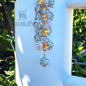 May include: A white Stanley tumbler with a decorative beaded charm. The charm features a cluster of clear, yellow, and iridescent beads, suspended from a silver-toned clasp. The tumbler has a handle and the Stanley logo.