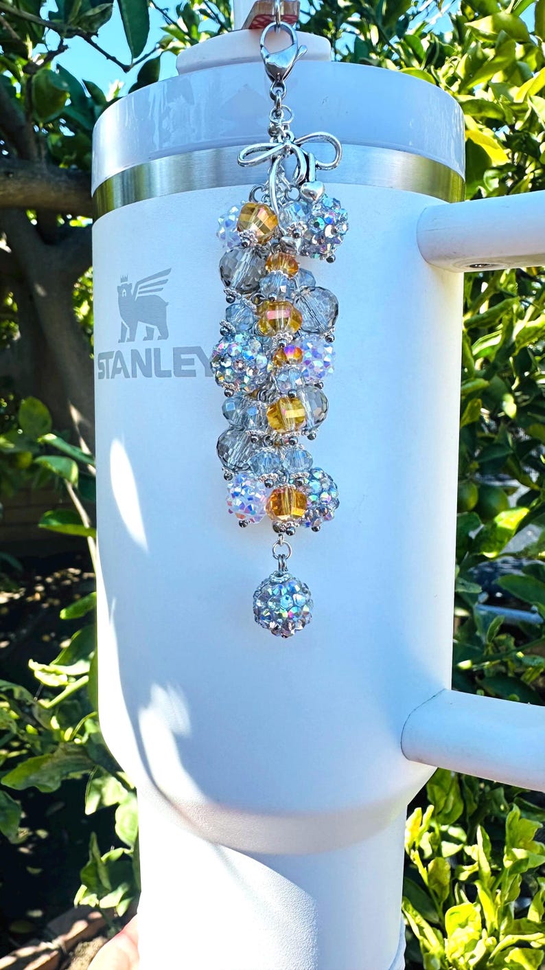 May include: A white Stanley tumbler with a decorative beaded charm. The charm features a cluster of clear, yellow, and iridescent beads, suspended from a silver-toned clasp. The tumbler has a handle and the Stanley logo.