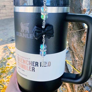 May include: Black Stanley Quencher 2.0 tumbler with a black handle and lid. The tumbler has a white band with the text "Quencher 2.0 Tumbler". A decorative charm with a black bow and iridescent beads hangs from the lid.