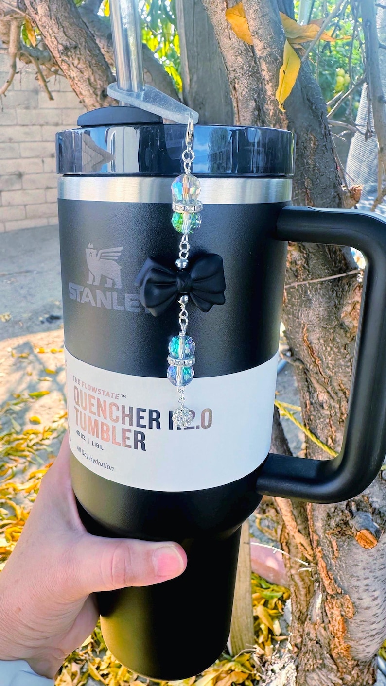 May include: Black Stanley Quencher 2.0 tumbler with a black handle and lid. The tumbler has a white band with the text "Quencher 2.0 Tumbler". A decorative charm with a black bow and iridescent beads hangs from the lid.