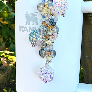 May include: A white Stanley tumbler with a decorative beaded charm. The charm features iridescent crystal beads in various sizes and colors, including clear, yellow, blue, and pink. The charm is attached to a silver-colored clasp.