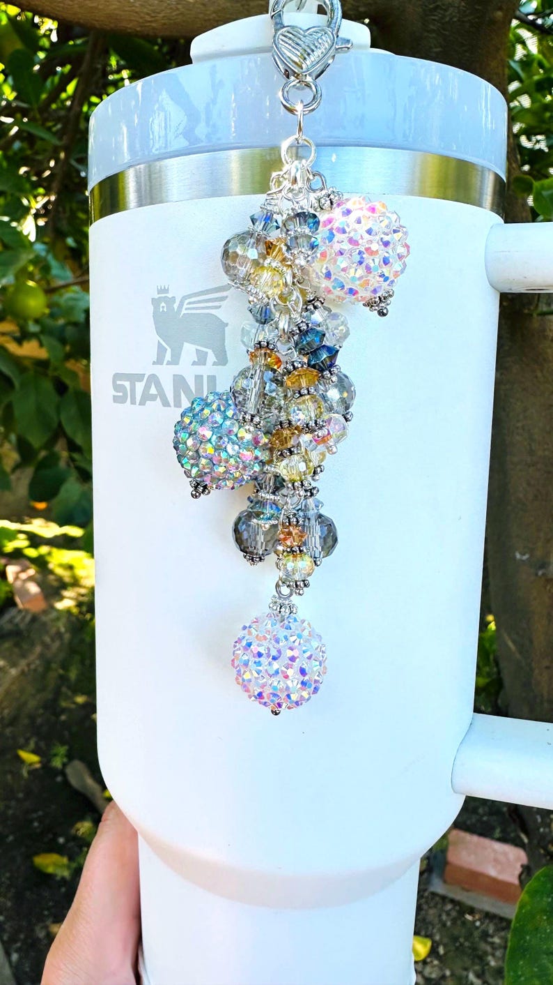 May include: A white Stanley tumbler with a decorative beaded charm. The charm features iridescent crystal beads in various sizes and colors, including clear, yellow, blue, and pink. The charm is attached to a silver-colored clasp.
