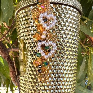 May include: A gold-colored tumbler with a textured, studded surface. The tumbler is adorned with a decorative cluster of pearls, gold beads, and colorful gems. A gold straw is inserted into the lid. The overall theme is luxury and glamour.