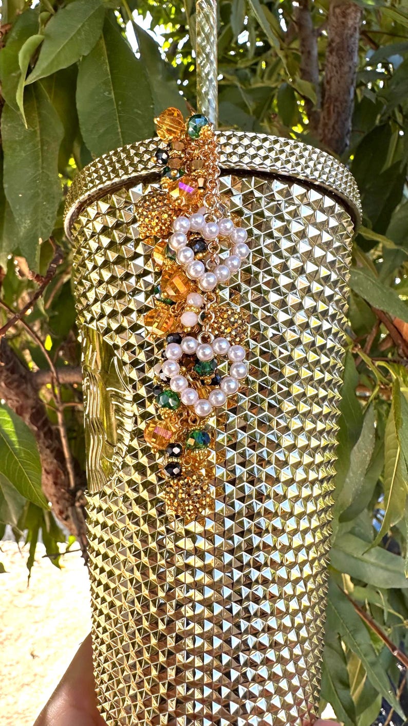 May include: A gold-colored tumbler with a textured, studded surface. The tumbler is adorned with a decorative cluster of pearls, gold beads, and colorful gems. A gold straw is inserted into the lid. The overall theme is luxury and glamour.