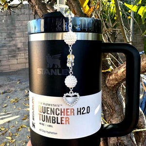 May include: Black 40 oz Stanley Quencher H2.0 tumbler with a handle. The tumbler has a white band with the text "QUENCHER H2.0 TUMBLER" and a decorative charm with crystal beads and a heart. The tumbler is designed for all-day hydration.