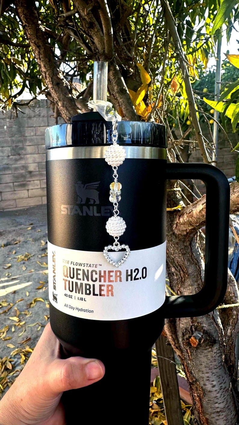 May include: Black 40 oz Stanley Quencher H2.0 tumbler with a handle. The tumbler has a white band with the text "QUENCHER H2.0 TUMBLER" and a decorative charm with crystal beads and a heart. The tumbler is designed for all-day hydration.