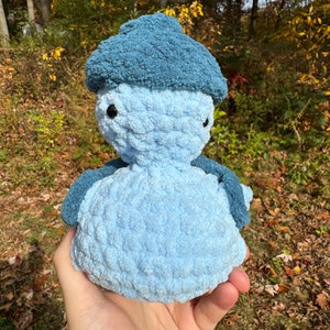 May include: A blue crocheted stuffed animal wearing a blue crocheted hat. The animal has a round body and a small, round head with two black eyes.