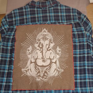 May include: A blue and white plaid flannel shirt with a brown patch on the back featuring a white image of the Hindu deity Ganesha.