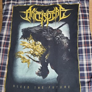 May include: A blue and white plaid flannel shirt with a large black graphic on the back. The graphic features a monster with the text "ARCHSPIRE" in gold and the words "BLEED THE FUTURE" in white.