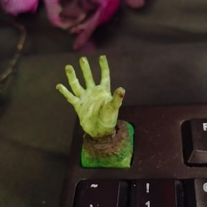 May include: A green, hand-shaped keyboard keycap. The hand is emerging from a green and brown base, with a small amount of red on one finger. The keycap is placed on a black keyboard.
