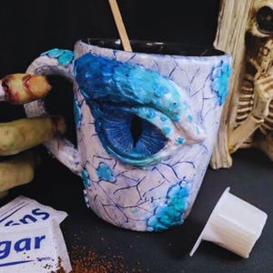 May include: A white ceramic mug with a blue and turquoise dragon eye design. The mug is filled with a dark liquid and a wooden stirrer is in the mug. There is a single-serve coffee pod and a packet of sugar next to the mug.