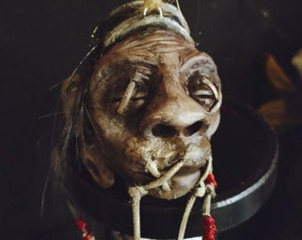 May include: A detailed, handcrafted sculpture of a human head with closed eyes, featuring intricate details and a weathered appearance. The head is suspended by twine and adorned with red beads and a feather, suggesting a tribal or ritualistic theme.