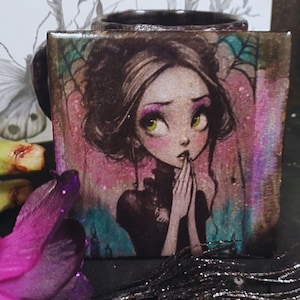 Puede incluir: Taza de cerámica cuadrada con una ilustración de una joven con ojos verdes, rubor rosa y cabello oscuro. El fondo tiene tonos rosas, turquesas y marrones. La taza tiene un asa y un borde negros.