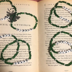 May include: Green beaded bracelets with white letter beads spelling out names like "Tom Riddle" and "Severus Snape" are displayed on an open book. The bracelets are a dark green color and the book pages are yellowed.