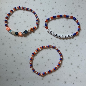 May include: Three beaded bracelets, each featuring a combination of orange, blue, and clear beads. Two bracelets spell out "ISLANDERS" in white letter beads. The bracelets are displayed on a white background with a star pattern.