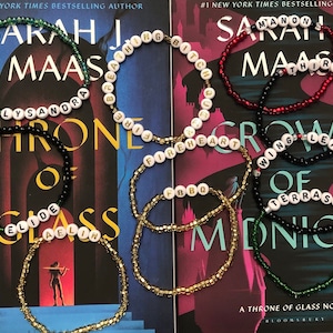 Puede incluir: Una colección de pulseras de cuentas de colores con cuentas de letras blancas que deletrean nombres y frases de la serie de libros "Throne of Glass" de Sarah J. Maas. Las pulseras se muestran en dos copias del libro, "Throne of Glass" y "A Throne of Glass Novel", de Sarah J. Maas.