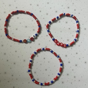 Könnte beinhalten: Drei Armbänder aus roten, weißen und blauen Perlen. Zwei Armbänder buchstabieren mit Buchstabenperlen "HABS". Die Armbänder liegen auf einer weißen Oberfläche mit Schneeflockenmuster.