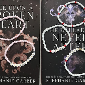 May include: Two stacks of books with beaded bracelets. One stack has two pink bracelets with the name "Evangeline" spelled out. The other stack has three white bracelets with the words "Jacks" and "Prince of Hearts".