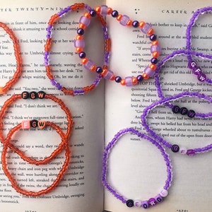 Puede incluir: Pulseras de cuentas en tonos naranja y morado, con letras que deletrean "FRED", "FGW" y "GEORGE". Las pulseras están sobre un libro abierto, mostrando un tema literario.