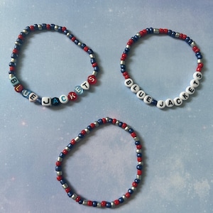 Può includere: Tre braccialetti di perline. Due braccialetti scrivono "BLUE JACKETS" con perline di lettere bianche. Gli altri braccialetti hanno perline rosse, blu e argento. I braccialetti sono su uno sfondo azzurro.