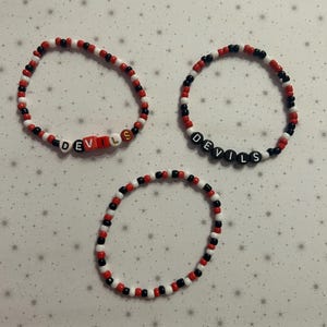 May include: Three beaded bracelets in red, black, and white. Two bracelets spell out "DEVILS" with letter beads. The bracelets are on a light gray background with a star pattern.