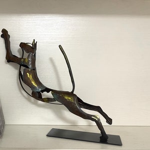 Op de afbeelding: Abstracte metalen sculptuur van een gestileerd dier, mogelijk een hond of een kat, dat met opgeheven staart naar voren springt. De sculptuur is gemaakt van metaal met een bruine, gouden en groene patina. Het is gemonteerd op een zwarte metalen voet.
