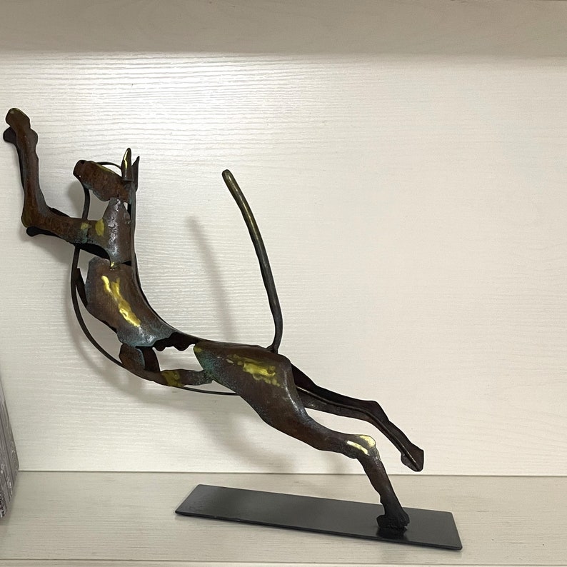 Op de afbeelding: Abstracte metalen sculptuur van een gestileerd dier, mogelijk een hond of een kat, dat met opgeheven staart naar voren springt. De sculptuur is gemaakt van metaal met een bruine, gouden en groene patina. Het is gemonteerd op een zwarte metalen voet.