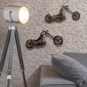 May include: Two metal motorcycle wall sculptures in a living room setting. The motorcycles are in a distressed, rusty finish with black accents. The motorcycles are mounted on a white brick wall. A gray couch with a gray pillow is in the foreground. A white tripod lamp with a white shade is in the background.
