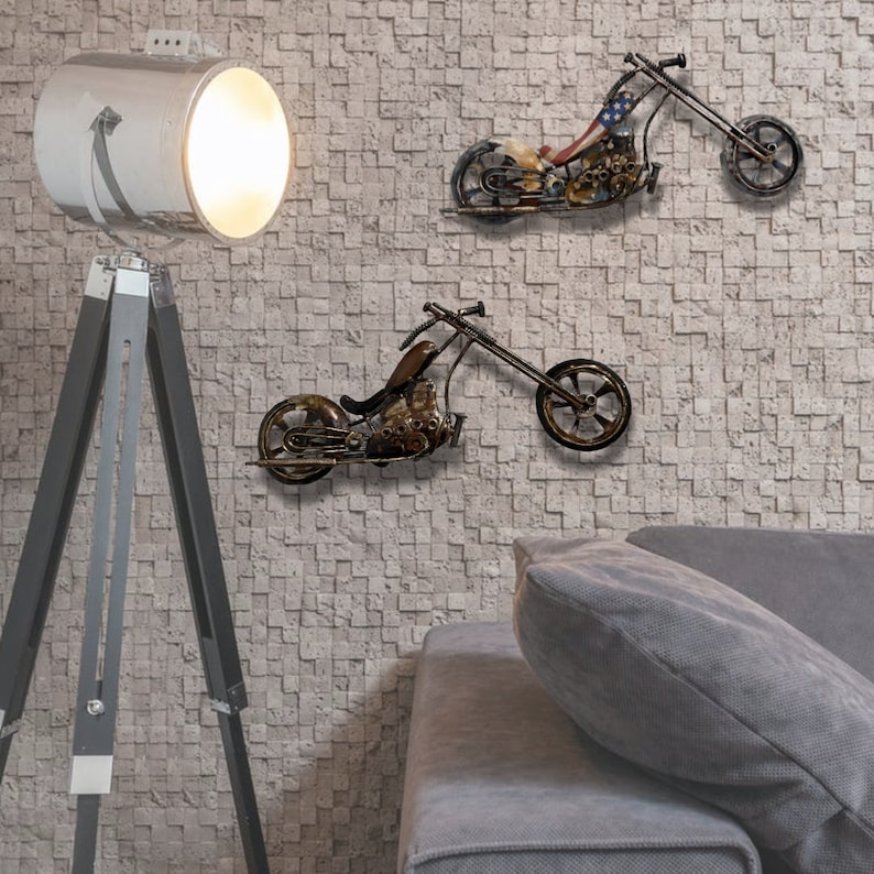 May include: Two metal motorcycle wall sculptures in a living room setting. The motorcycles are in a distressed, rusty finish with black accents. The motorcycles are mounted on a white brick wall. A gray couch with a gray pillow is in the foreground. A white tripod lamp with a white shade is in the background.