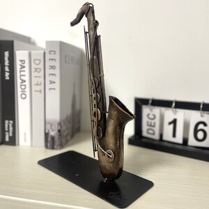 May include: A metal saxophone sculpture with a brown and gold finish. The sculpture is on a black stand and is in front of a white wall.
