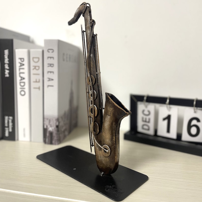 May include: A metal saxophone sculpture with a brown and gold finish. The sculpture is on a black stand and is in front of a white wall.