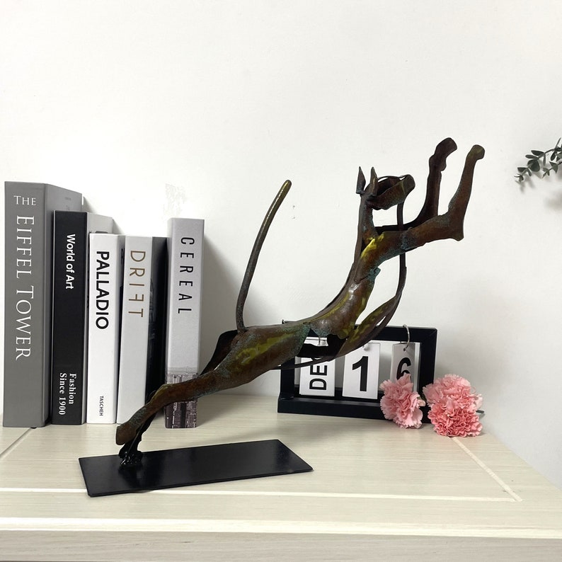 Op de afbeelding: Een bronzen metalen sculptuur van een springende kat. Het beeld staat op een zwarte basis en wordt tentoongesteld op een tafel met boeken en een kalender.