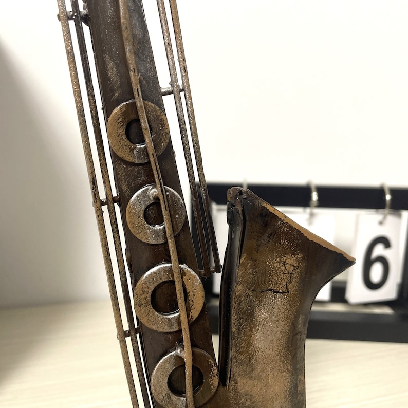 May include: A metal saxophone sculpture with a distressed brown and silver finish. The saxophone has a bell-shaped mouthpiece and is decorated with decorative rings.