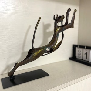 Op de afbeelding: Een bronzen metalen sculptuur van een springende kat met een lange staart. Het beeld is gemonteerd op een zwarte basis.