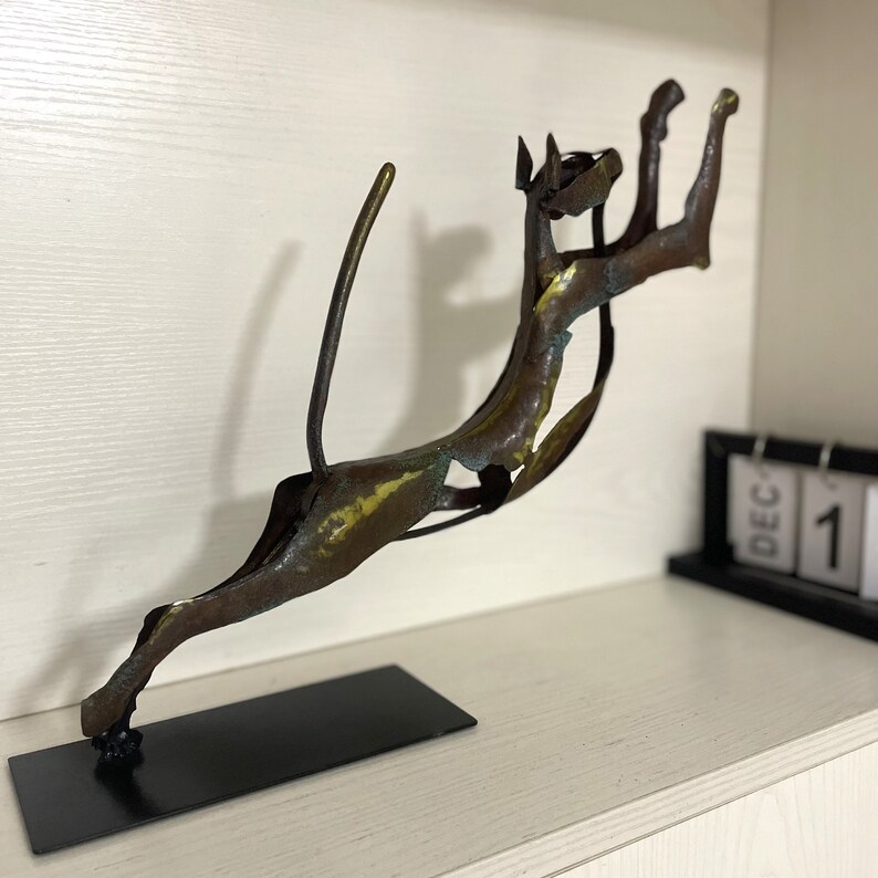 Op de afbeelding: Een bronzen metalen sculptuur van een springende kat met een lange staart. Het beeld is gemonteerd op een zwarte basis.
