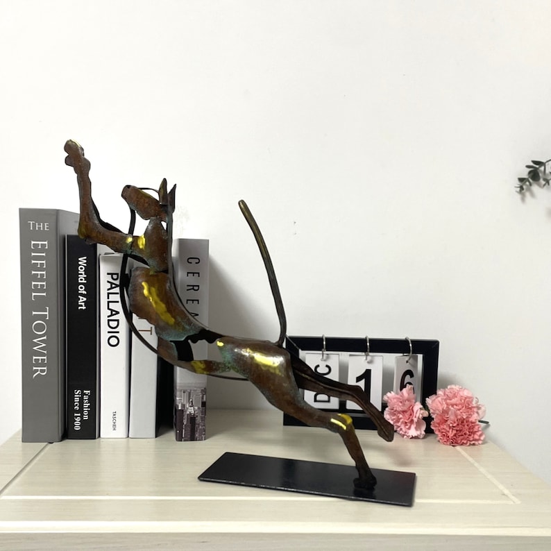 Op de afbeelding: Een bronzen metalen sculptuur van een springende kat. Het beeld staat op een zwarte basis en wordt tentoongesteld op een tafel met boeken en een kalender.